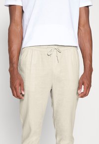 Homme portant un pantalon beige à cordon de serrage et une chemise blanche à manches courtes, montré de la moitié de la poitrine à la moitié des cuisses sur un fond uni.