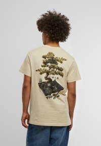 Beige cotton t-shirt featuring a graphic design of a bonsai tree on a turntable with the text "The Root of all." Short sleeves, relaxed fit.