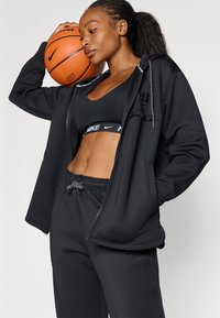 Conjunto deportivo negro que incluye una chaqueta con capucha y cremallera, top corto y pantalones elásticos con cordón; textura suave y diseño ajustado.