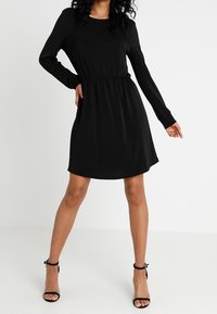 Svart långärmad klänning med en figursydd midja och flödande kjol. Tillverkad av mjukt tyg, med rund urringning och enkel design.