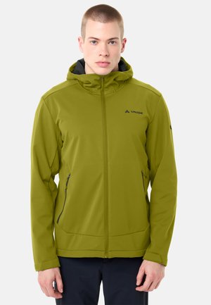 Giovane uomo che indossa una giacca con cappuccio verde oliva con cerniera frontale e tasche laterali con cerniera, in piedi contro uno sfondo chiaro e uniforme.