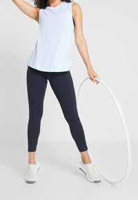 Light blue sleeveless top paired with navy leggings. Model holds a white hula hoop. Footwear is white with gray accents. Smooth texture throughout.