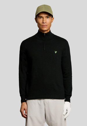Mand iført en sort quarter-zip sweater med en grøn logo, beige bukser, en beige perforeret kasket og en hvid golfhandske på sin højre hånd.