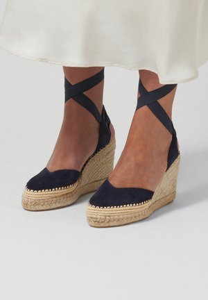 Wedges met marineblauwe suède gesloten tenen, plateauhakken van gevlochten jute, en zwarte enkelbandjes, gedragen met een witte rok.