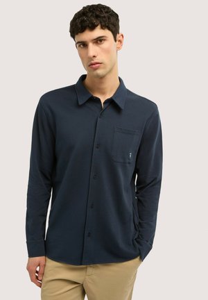 Joven con camisa azul marino de manga larga con botones y bolsillo en el pecho, y pantalones beige, de pie frente a un fondo claro liso.