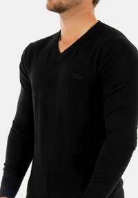 Mann trägt einen schmal geschnittenen schwarzen V-Ausschnitt-Pullover mit langen Ärmeln und dezentem Logo auf der Brust sowie einer dünnen goldenen Kette.