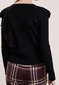 Schwarzer gerippter Pullover mit einer großen Rüschendetail entlang des Rückens, kombiniert mit Karo-Hosen in burgunderfarbenen, cremefarbenen und schwarzen Tönen.