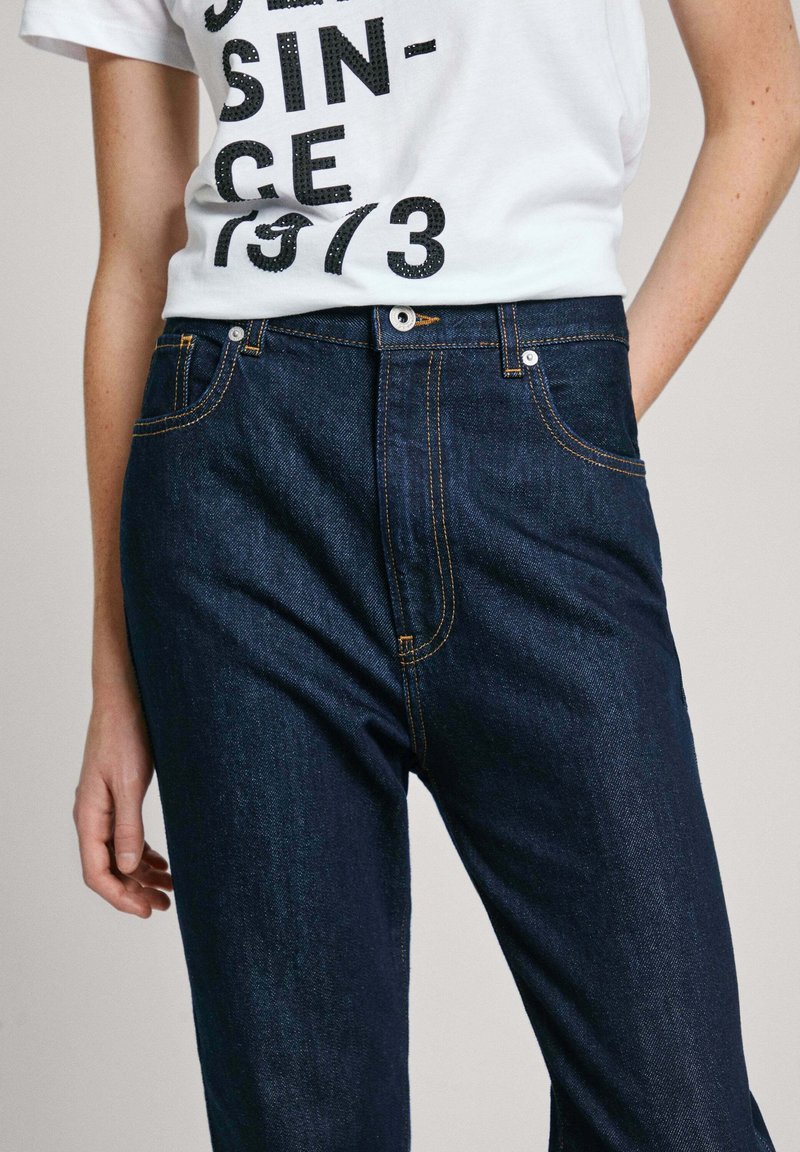 Dunkelblaue Jeans aus Denim mit orangefarbenen Nähten, einem klassischen Fünf-Taschen-Design und einem frontalen Knopfverschluss. Glatte Textur und eng anliegende Form.