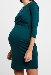 Elastyczna turkusowa sukienka ciążowa z zawiązanym przodem, z r�ękawami do 3/4 i dopasowanym krojem podkreślającym brzuszek.