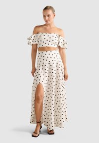 Žena v bielom off-shoulder crop top s vysokým pásom, zladená s sukňou s bodkami a rozparkom na stehne, doplnená čiernymi sandálmi na podpätku.