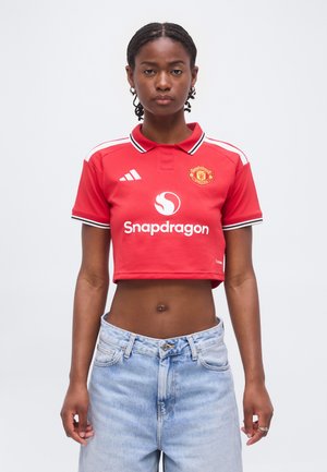 Kvinde iført en rød Manchester United cropped polo med Snapdragon-logo og lyseblå højtaljede jeans, står mod en ensfarvet baggrund.