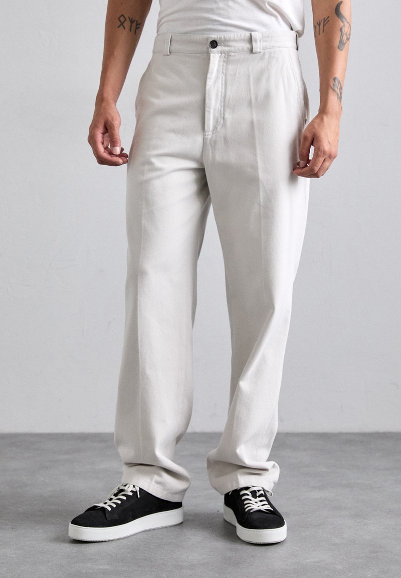 Man wearing loose light beige pants and black sneakers with white soles standing on gray concrete floor.