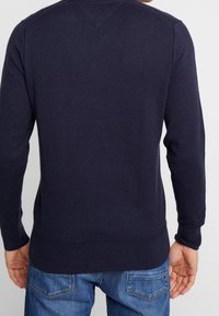 Granatowy sweter dziany z długimi rękawami, ribbowanymi mankietami i dołem, oraz detalem w kształcie litery V na szyi, połączony z dżinsami, widok od tyłu.