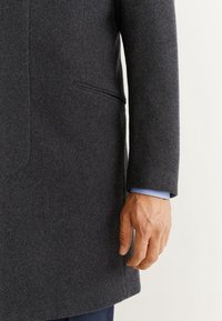 Abrigo de lana gris con corte recto, que presenta un bolsillo lateral y un puño de camisa azul visible por debajo de la manga.