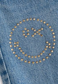 Visage souriant composé de petits clous dorés sur un tissu en denim bleu, avec des yeux en forme de X et une bouche incurvée.