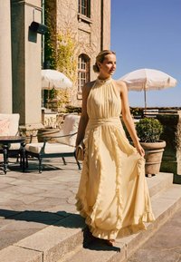 Femme en robe fluide jaune pâle descendant des marches en pierre à l'extérieur d'un bâtiment ensoleillé avec des sièges extérieurs et des parasols.