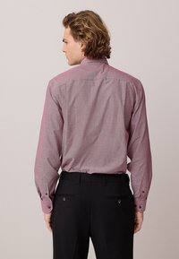 Uomo con capelli mossi che indossa una camicia rosa a maniche lunghe con bottoni e motivo, infilata in pantaloni eleganti neri, visto di spalle su sfondo neutro.