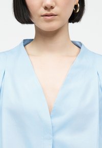 Femme aux cheveux courts et foncés portant un chemisier bleu ciel à col en V et des boucles d'oreilles en forme de créoles dorées, sur un fond clair uni.