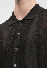 Μαύρο πόλο μπλουζάκι με υφή πλεκτής σχεδίασης, που διαθέτει κάθετες και οριζόντιες γραμμές, εξοπλισμένο με γιακά και κλείσιμο με κουμπί.