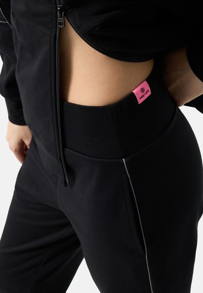 Close view of a person wearing black zip-up top and black pants with wide waistband and pink Fire+Ice label on the hip.