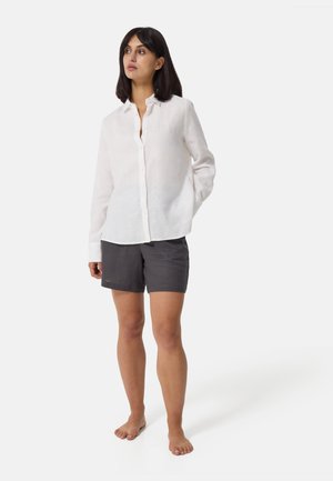 Hvid linned skjorte med lange ærmer og knaplukning, afslappet fit, parret med mørkegrå linnedshorts. Modellen står barfodet med den ene hånd i lommen.