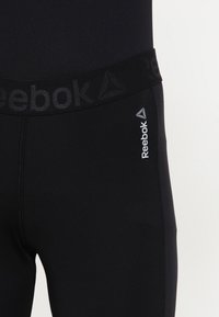 Czarne legginsy Reebok z szerokim elastycznym pasem, na którym znajduje się logo marki. Materiał jest gładki, o dopasowanym kroju i płaskich szwach.