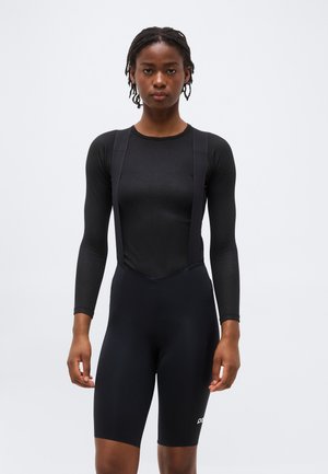 Femme aux cheveux tressés portant un haut noir à manches longues et un cuissard de cyclisme noir, debout devant un fond uni.