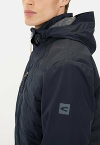 Navy nylon jacket features a quilted texture, adjustable hood, zippered chest pocket, and logo detail on the left sleeve.
