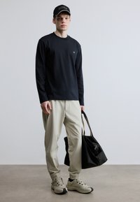 Fekete hosszú ujjú ing, krémszínű szűkített nadrág, bézs sportcipők és egy fekete fogantyús táska. A sapkán fehér "BOSS" hímzett logó található.