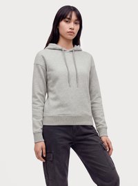 Junge Person mit langen schwarzen Haaren, die einen hellgrauen Hoodie und schwarze Cargohosen trägt und vor einem schlichten Hintergrund steht.