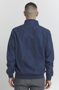 Marineblå vindjakke med opretstående krave, elastiske manchetter og kant. Glat tekstur, ensfarvet, minimal dekoration bagpå.