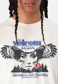 White cotton t-shirt featuring a graphic with an owl, a woman's face, and text in blue, black, and red. Includes forest silhouette details.