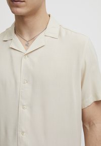 Kortermet beige skjorte med krage, med en glatt tekstur og subtil glans, knappet front og avslappet passform. Sølvkjede synlig.