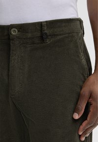 Donkergroene corduroy broek met verticale ribbels, voorzien van een knop en een vierkante zwarte accent. De voorzakken zijn zichtbaar aan de zijkanten.