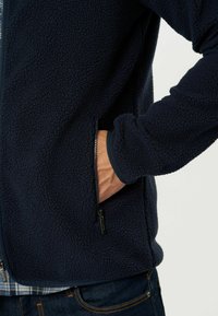 Tumedast fleece'ist jakist, millel on pehme ja tekstureeritud viimistlus, funktsionaalne lukuga tasku vööl ja soonikkoes varrukaotsad kindla sobivuse tagamiseks.