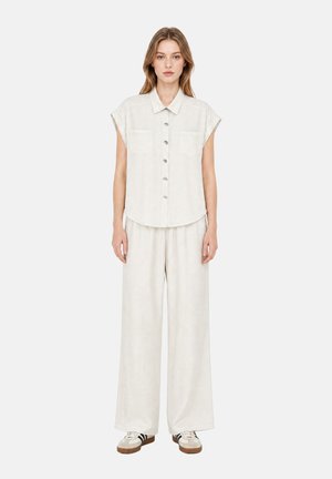 Jeune femme portant une chemise blanche boutonnée à manches courtes ample et un pantalon blanc à jambes larges avec des baskets blanches sur un fond uni.