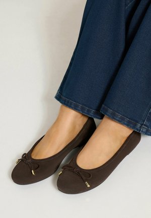 Pieds portant des ballerines marron foncé avec de petits nœuds, assortis à un jean en denim bleu.