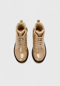 Une paire de bottes en or à paillettes avec des lacets, doublées de fourrure et à semelles noires, vue de dessus sur un fond blanc.