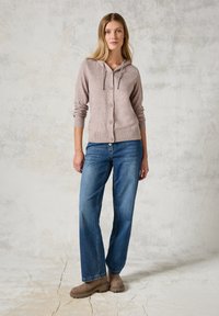 Cardigan à capuche marron clair avec des boutons et des poignets côtelés, associé à un jean bleu à jambe large et des bottines beiges. Design minimaliste.