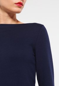 Tummansininen pitkähihainen paita, jossa on venepääntie, valmistettu sileästä ja kevyestä kankaasta. Ominaisuuksiin kuuluu tiukat hihat ja minimalistinen muotoilu.
