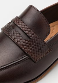 Mocassin en cuir marron avec une bande tressée en cuir, texture lisse et coutures détaillées ; bout arrondi et semelle antidérapante.