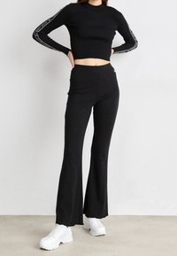 Černý žebrovaný crop top s dlouhým rukávem a logem na rukávech, v kombinaci s černými rozšířenými kalhotami a bílými chunky teniskami.