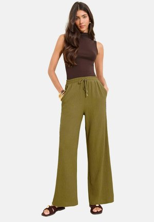 Kobieta z długimi ciemnymi włosami, ubrana w czarny top bez rękawów, oliwkowozielone szerokie spodnie ze sznurkiem i brązowe sandały, stojąca z rękami w kieszeniach.