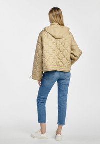 Beżowa pikowana kurtka puchowa z kapturem, z wzorzystą teksturą i oversize'owym krojem, noszona na jasnoniebieskie dżinsy i białe sneakersy.