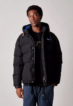 Giovane uomo che indossa una giacca a vento nera con cappuccio e interno blu, pantaloni blu navy e una maglietta nera a collo rotondo con stampa grafica.