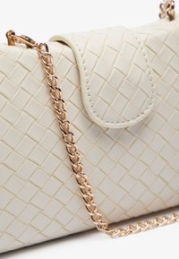 Sac à main en crème tissé avec une fermeture à rabat et une bandoulière en chaîne dorée rose. Il présente une surface texturée avec des motifs en diamant et des détails de couture.