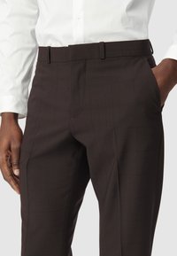 Pantalons en flanelle à carreaux marron foncé avec une texture de tissu lisse, dotés de passants de ceinture et de poches latérales, complétés par une chemise blanche.