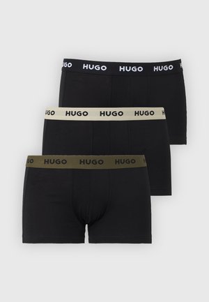 Tall, muscular man stands barefoot in black HUGO boxer briefs, arms behind back, gazing to the right against plain white background.