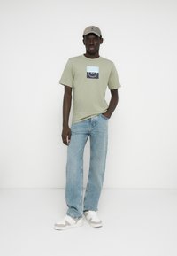 Ανοιχτό πράσινο βαμβακερό μπλουζάκι με τυπωμένο γραφικό, συνδυασμένο με ανοιχτό μπλε τζιν και λευκά αθλητικά παπούτσια με γκρι λεπτομέρειες και μπεζ καπέλο.