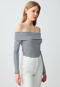 Ein schulterfreies, geripptes Oberteil mit navyblauen und weißen Streifen, langen Ärmeln und figurbetonendem Schnitt, kombiniert mit weißen Jeans.